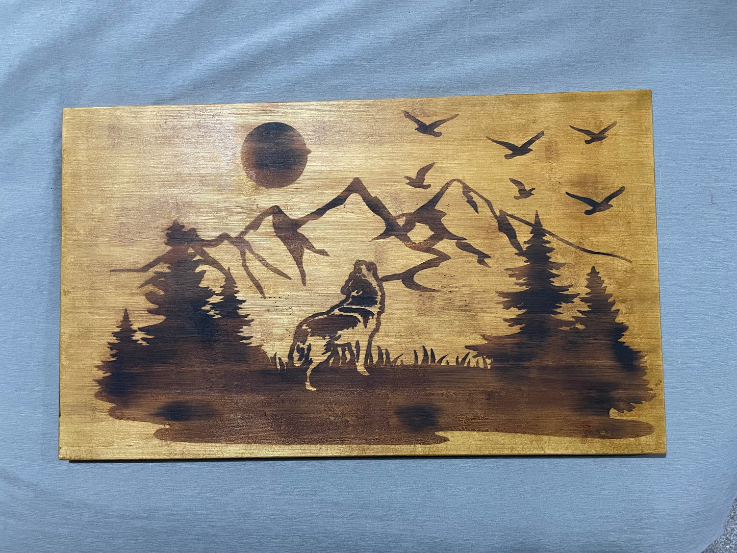 Wooden plaque with burnt landscape of a wolf, trees, mountains, and birds.